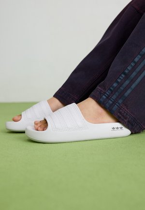 Pieds portant des claquettes blanches Adidas avec des brides texturées, reposant sur une surface verte, accompagnés d'un pantalon foncé à trois bandes.