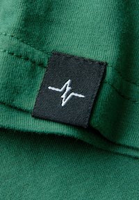 Grüner Baumwollstoff mit sichtbaren Nähten. Verfügt über ein schwarzes Etikett mit einem weißen Herzschlag-Design. Weiche Textur, lässiges Design.