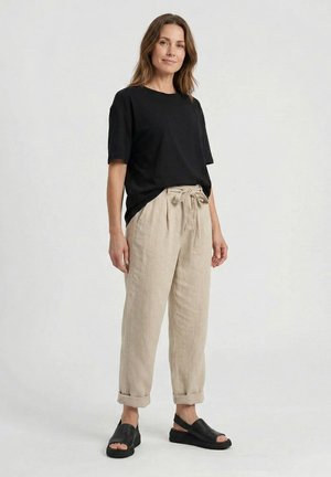 Kvinde stående iført en løs sort T-shirt, beige høj talje bindebukser med op-rullede manchetter og sorte åbne tå-sandaler på en ensartet baggrund.