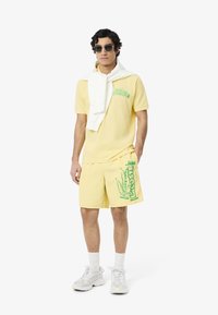 Polo giallo con logo verde, pantaloni shorts gialli abbinati con grande stampa verde, maglione bianco drappeggiato sulle spalle, sneaker grigie.