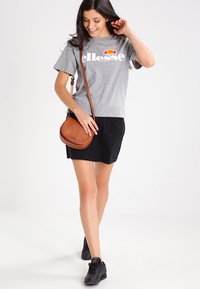 Sivi kratki rukav t-shirt s bijelim "ellesse" logotipom, crna A-linija suknja, smeđa torba preko tijela i crne tenisice. Glatka tekstura tkanine.