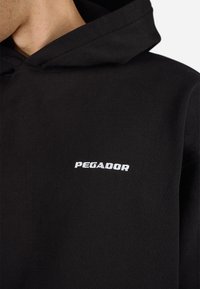 Svart hettegensere laget av bomull, med en frontlomme i kenguru-stil og en hvit brodert "PEGADOR" logo på brystet.
