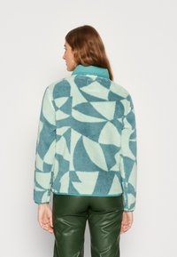 Fleecejack in licht teal met abstracte geometrische patronen in donkerder teal. Heeft een contrasterende kraag en een zachte textuur.