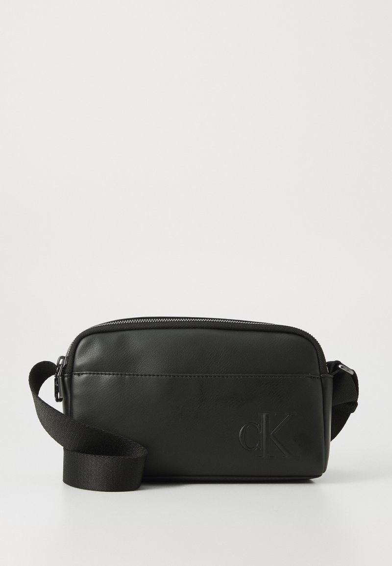 Sac en cuir noir avec fermeture éclair, bandoulière réglable et logo en relief. Forme rectangulaire, texture lisse, design minimal.