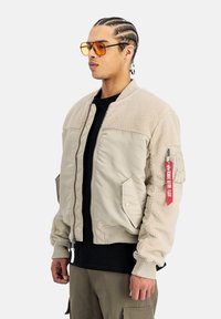 Chaqueta bomber beige con parte superior de forro polar, bolsillos con cremallera, puños de canalé y una etiqueta roja en la manga. Usada con una camiseta negra debajo.