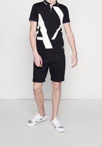 Svart polo t-shirt med en vit geometrisk design, ihopparad med svarta shorts och rutig vita sneakers. Enkel krage och skräddarsydd passform.