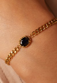 Gouden kettingarmband met een ovale zwarte steen als middelpunt, met ingewikkelde details rond de steen en gladde schakels.