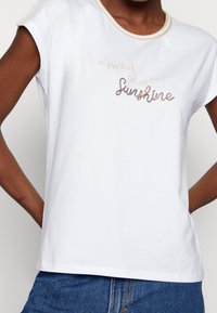 Hvid bomulds T-shirt med korte ærmer, prydet med metalisk guld skrift, der lyder "a pocket full of Sunshine", samt et lille lommegrafik.