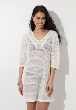 Donna con capelli lunghi e scuri che indossa un abito bianco all'uncinetto sopra biancheria nera, sorridente contro uno sfondo chiaro e semplice.