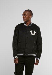 Must tähnikas must varsity jacket valgete triipudega krael ja varrukaotsadel, silmapaistva logo plaastri ja kahe küljetaskuga.