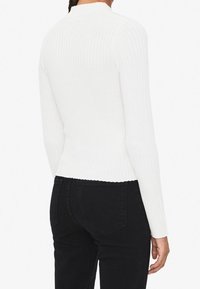 Haut blanc à manches longues côtelé avec un col montant, coupe ajustée et tissu texturé. Porté avec un pantalon noir taille haute, affichant un style minimaliste.