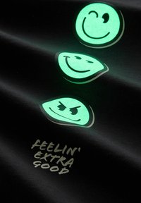 Tela negra con tres diseños de caras sonrientes que brillan en la oscuridad en verde. Incluye el texto "FEELIN' EXTRA GOOD" en gris claro debajo de los emojis.