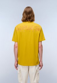 Person with shoulder-length hair wearing a mustard yellow T-shirt with faded text and off-white cargo pants, facing away against a plain background.