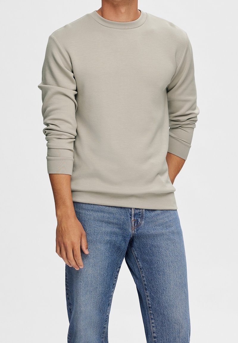 Sudadera beige clara de mezcla de algodón, con cuello redondo, mangas largas y puños acanalados, combinada con vaqueros azules.