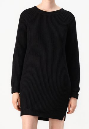 Femme portant une robe pull noire côtelée à manches longues, avec un col rond et une petite fente latérale à l'ourlet.