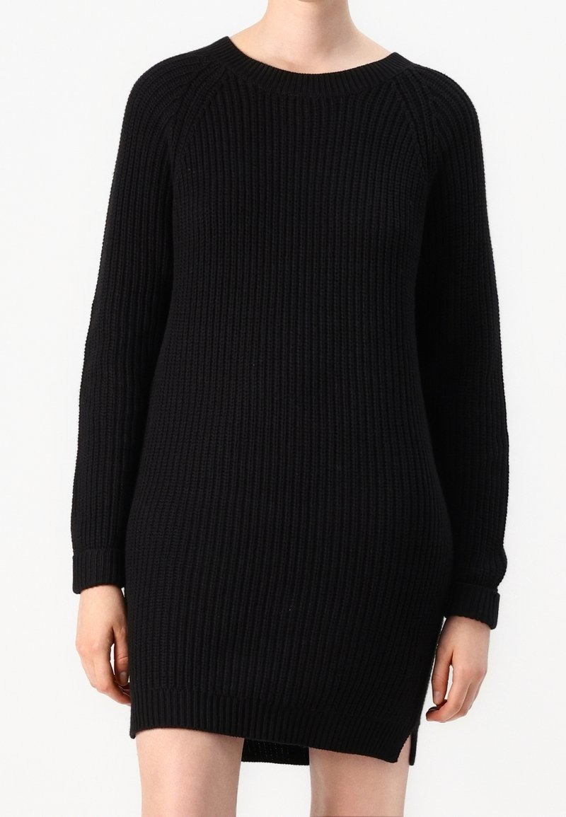 Femme portant une robe pull noire côtelée à manches longues, avec un col rond et une petite fente latérale à l'ourlet.