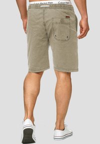 Licht khaki short met een elastische tailleband, zij- en achterzakken, en een textuurstofafwerking. Witte logoband zichtbaar.