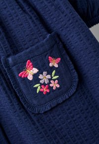 Marinefarbener strukturierten Stoff mit einer Tasche, die mit bunten gestickten Blumen und Schmetterlingen verziert ist, akzentuiert durch einen volanten Rand.