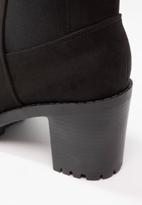 Schwarze Wildleder-Ankle-Boots mit gestapeltem Blockabsatzdesign und elastischem Seitenpaneel auf weißem Hintergrund.