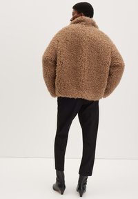 Giacca esterna marrone fluffy con colletto alto, vestibilità oversize e texture morbida. Abbinata a pantaloni neri su misura e stivali con tacco alla caviglia.