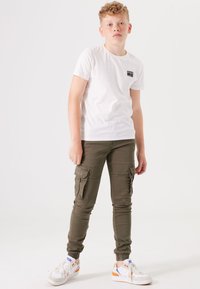 Weißes T-Shirt mit kleinem schwarzem Logo, olivfarbene Cargo-Shorts mit Seitentaschen und weiße Sneaker mit orangen Akzenten. Gepolsterte Knöchelabschlüsse.