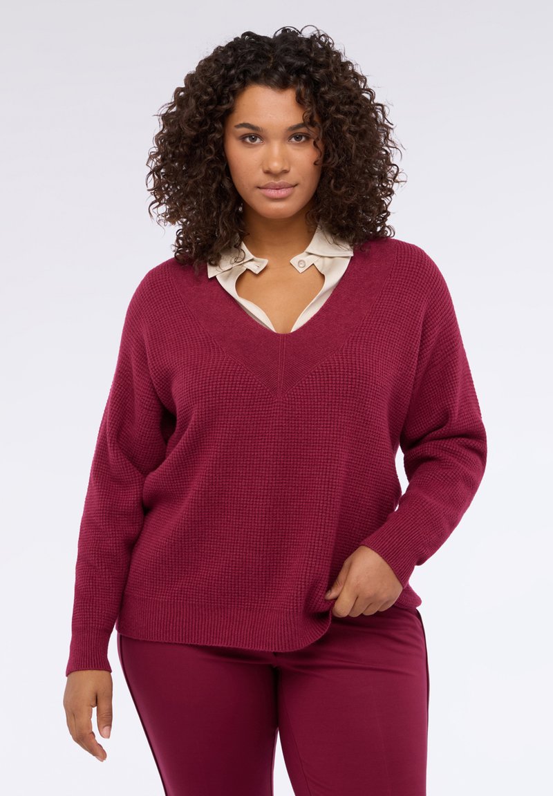 Maglione bordeaux lavorato a maglia con scollo a V, vestibilità ampia e tessuto strutturato. Abbinato a una camicia crema con colletto sotto.