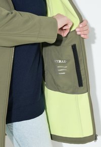 Voděodolná bunda vyrobená z khaki materiálu, s jasně limetkově zeleným interiérem a zipovou kapsou s štítky uvnitř.