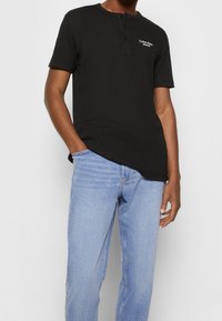 Man som bär en svart kortärmad henley-skjorta med "Calvin Klein Jeans"-logotyp och ljusblå denimjeans, med handen i fickan.