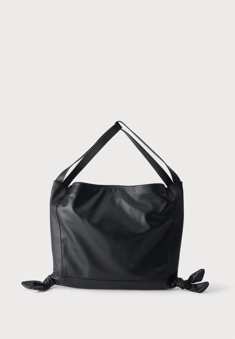 Sac d'épaule en cuir noir avec larges bretelles et détails noués à chaque coin inférieur sur un fond clair uni.