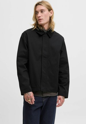 Black waterproof jacket with a buttoned collar, smooth texture, and a relaxed fit. Paired with dark jeans and visible tattoos on hands.
