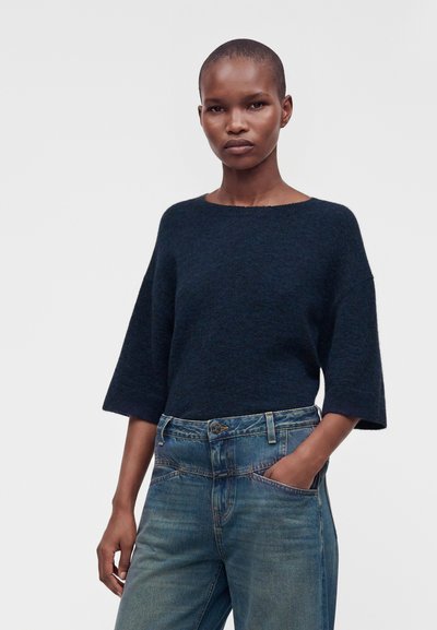 Femme aux cheveux courts portant un pull bleu foncé à manches trois-quarts et un jean bleu, une main dans la poche.