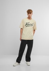 Beige t-shirt med kortærmer og grønt "Kani" logo, parret med mørkegrå bukser. Stoffet ser blødt og afslappet ud. Hvide sneakers fuldender outfittet.