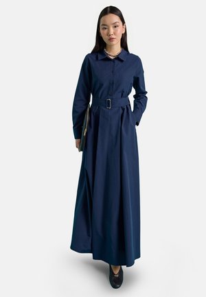 Donna che indossa un lungo vestito camicia blu navy con cintura, tiene una pochette in una mano, in piedi davanti a uno sfondo chiaro e uniforme.