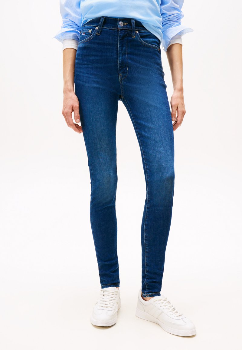 Person iført mørkeblå skinny jeans og hvide sneakers, med en lyseblå skjorte stoppet ind, stående mod en hvid baggrund.