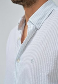 Camicia a righe blu chiaro e bianco, con una texture morbida, colletto standard e un piccolo logo sul petto.