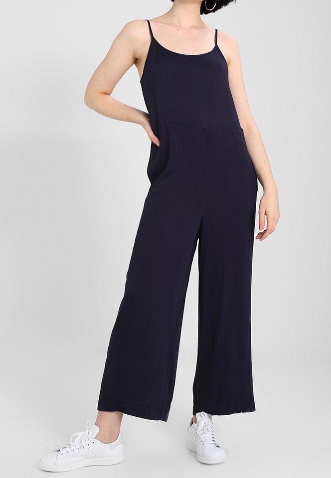 Femme portant une combinaison sans manches bleu marine à jambes larges et des baskets blanches, debout avec une main sur la hanche sur un fond uni.