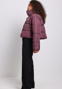 Chaqueta acolchada corta y ajustada en morado profundo con un cuello alto, con costuras horizontales y cierre de cremallera, combinada con pantalones negros.