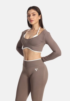 Žena v taupe crop top so zapínaním na dlhý rukáv a zodpovedajúcich legínach s vysokým pásom, stojaca s rukami na bokoch pred jednoduchým pozadím.