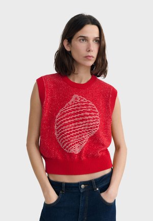 Femme portant un haut rouge sans manches en maille avec un motif central texturé et abstrait, associée à un jean bleu foncé, debout avec les mains dans les poches.