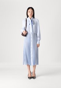 Blusa azul clara com colarinho laço, combinada com uma saia plissada azul clara com botões à frente. O modelo usa sapatos de salto alto pretos.