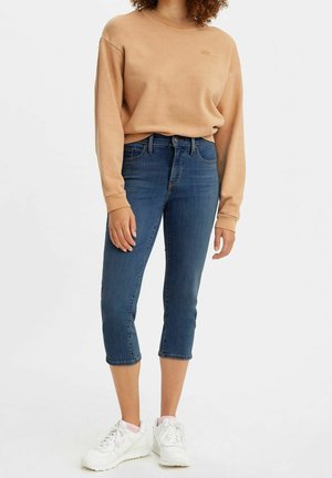 Persona que lleva una sudadera beige de manga larga, vaqueros azules recortados y zapatillas blancas, de pie frente a un fondo neutro.