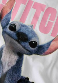 Cartoonfiguur met blauwe vacht, grote zwarte ogen en opvallende roze oren, tegen een witte achtergrond met het woord "STITCH" in roze.