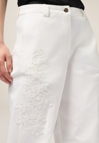 Witte katoenen broek met ingewikkelde witte bloemenborduurwerk aan de linkerkant, met een slank ontwerp en een klein knoopdetail aan de voorkant.