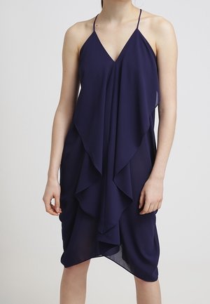 Robe bleu marine sans manches avec fines bretelles, volants superposés à l'avant, ourlet asymétrique, portée par une personne debout sur un fond uni.