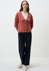 Cardigan rosa lavorato a maglia con texture a diamante, scollatura a V e cinque bottoni dorati. Abbinato a pantaloni larghi in denim scuro e sneakers bianche.