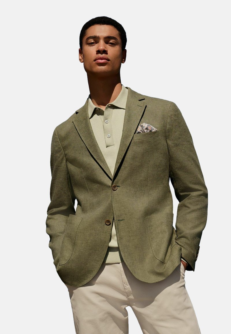 Olivgrüner Blazer mit zwei Knöpfen und Reverskragen, kombiniert mit einem hellgrünen Poloshirt. Enthält ein gemustertes Taschenquadrat und cremefarbene Hosen.