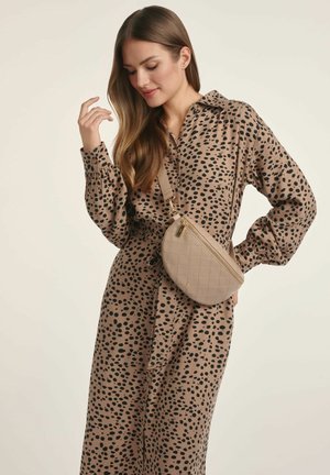 Beżowa sukienka koszulowa z czarnym nadrukiem w kropki, z długimi rękawami, kołnierzykiem i związanym w pasie. Mała pikowana torebka na ramię w jasnobeżowym odcieniu.