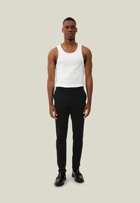 Bela brezrokavnik, kombiniran s črnimi hlačami slim fit in črnimi čevlji. Izgled se odlikuje po minimalističnem dizajnu in nevtralnem ozadju.
