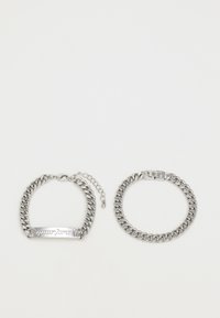 Silverkedjearmband med en platt stav med ingraverad text, en med en löstagbar krok och en justerbar förlängningskedja.