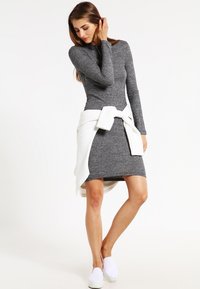 Grå strikket bodycon-kjole med lange ermer, rund hals og en hvit genser knyttet rundt livet. Brukt med hvite slip-on sko.